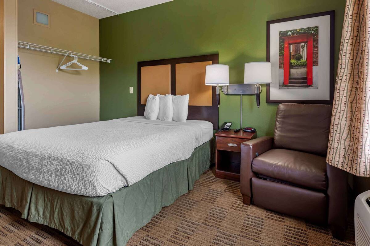 A hotel room with a bed, a chair, a nightstand, and a wall-mounted lamp.