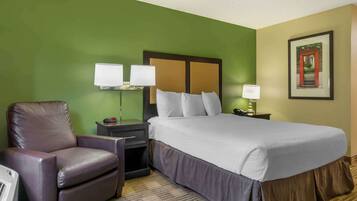 Una habitación de hotel con una cama grande, un sillón de cuero, una mesita de noche con lámpara y un cuadro en la pared.