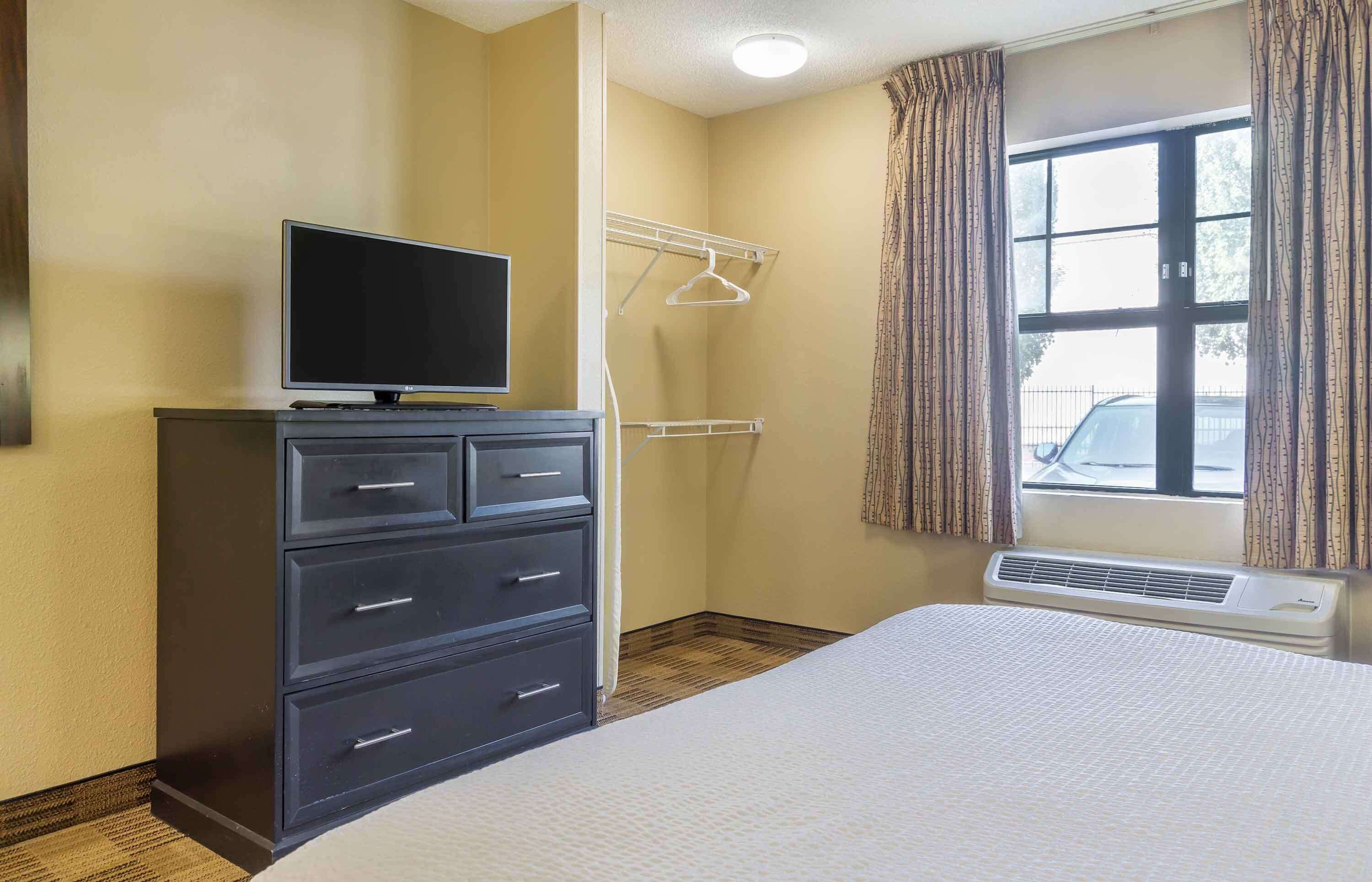 Une chambre d’hôtel avec un lit, un téléviseur posé sur une commode, une fenêtre avec des rideaux et un climatiseur fixé au mur.