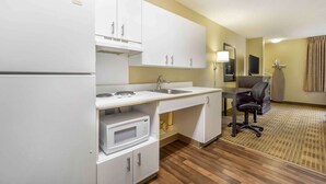 Cocina de hotel compacta con armarios blancos, microondas, lavamanos y un pequeño comedor con una silla.
