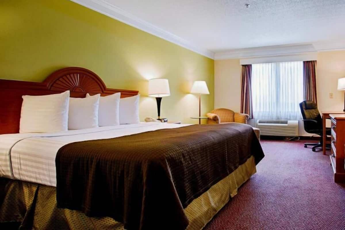 A hotel room with a large bed, two bedside lamps, a desk with a chair, an air conditioning unit, and a window with curtains.