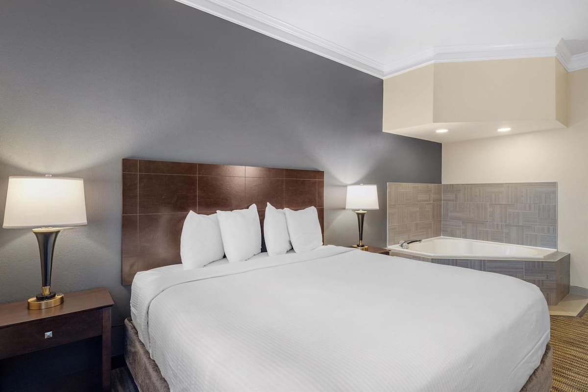 A hotel room with a large bed, two bedside lamps, a wooden headboard, a built-in bathtub, and a neutral color scheme.