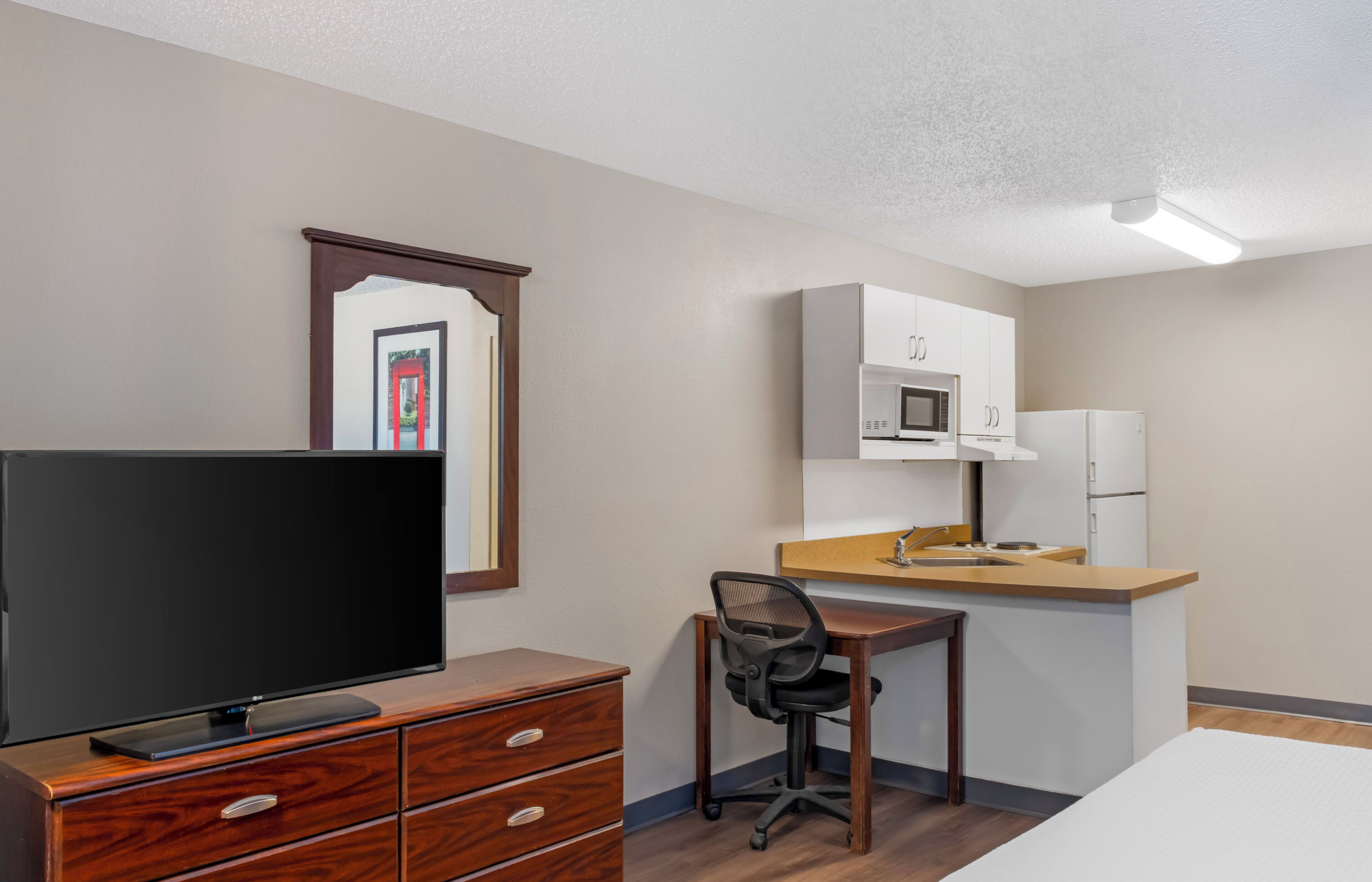 Quarto de hotel com televisão, cómoda de madeira, secretária, cadeira, kitchenette e espelho.