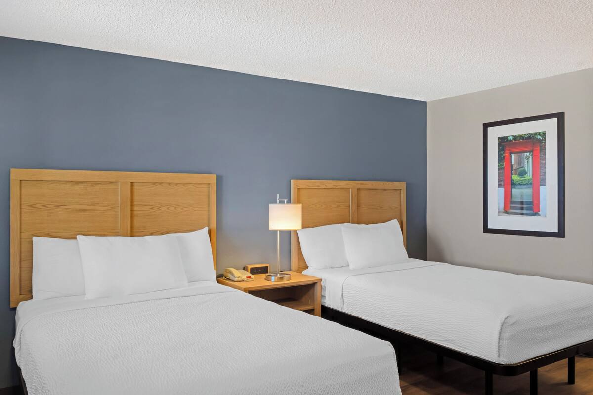 A hotel room with two single beds, wooden headboards, a nightstand with a lamp, a framed picture on the wall, and a telephone on the nightstand.