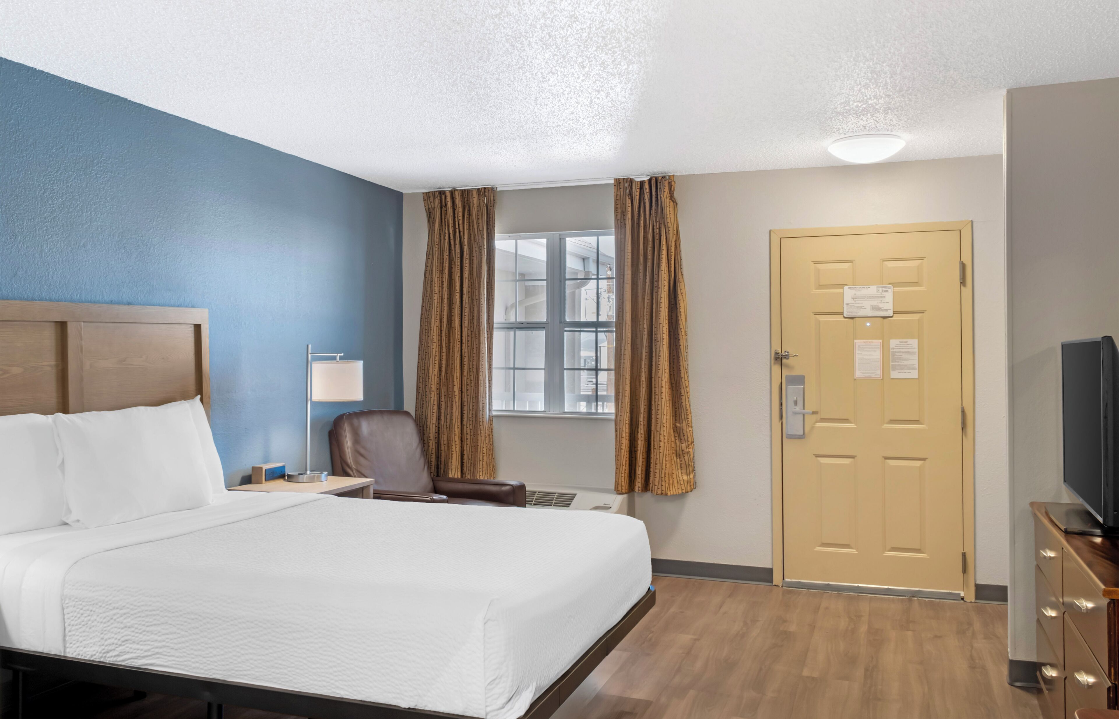 Habitación de hotel con cama, escritorio, silla, televisión, puerta y ventana con cortinas.