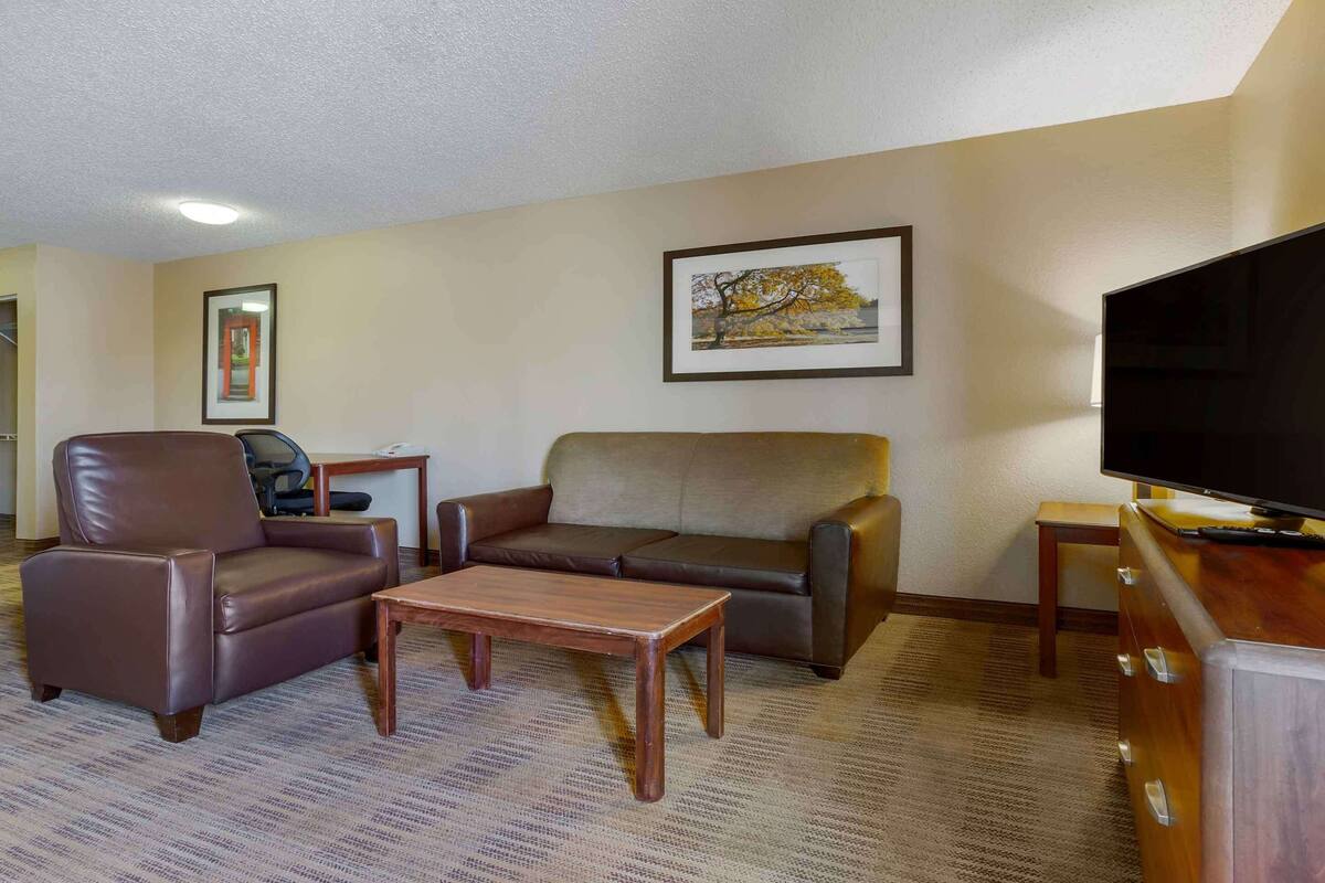 A hotel room with a brown leather sofa, a brown leather armchair, a wooden coffee table, a television on a wooden stand, and a framed picture on the wall.