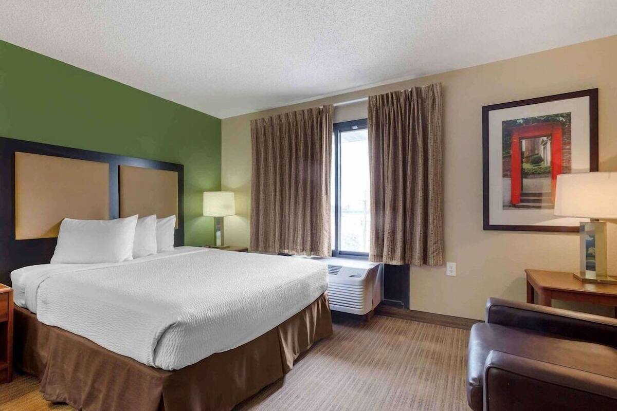 A hotel room with a bed, a desk, a chair, a window with curtains, an air conditioning unit, and a framed picture on the wall.