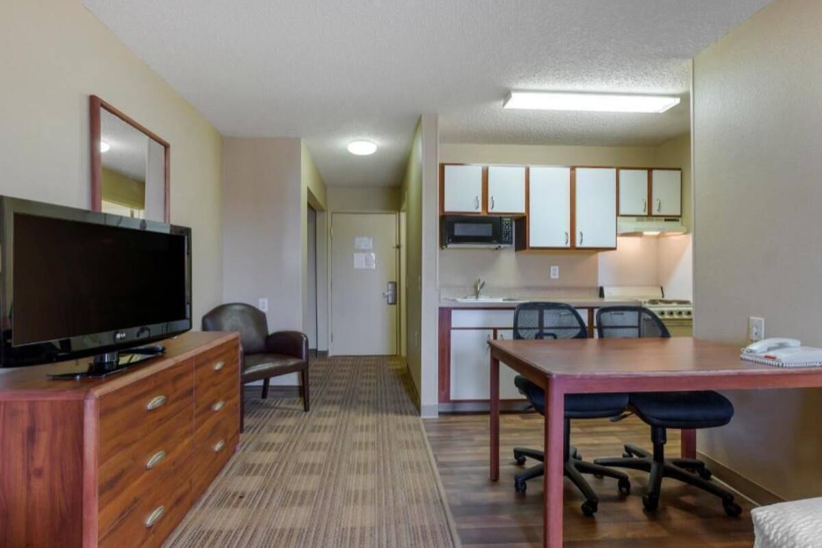 A hotel room with a TV, wooden dresser, carpeted floor, kitchenette, and a dining area with chairs.
