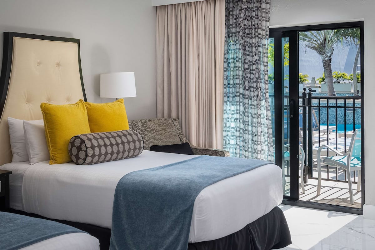 A hotel room with a bed, a nightstand, a lamp, a view of a pool and palm trees through a sliding glass door.