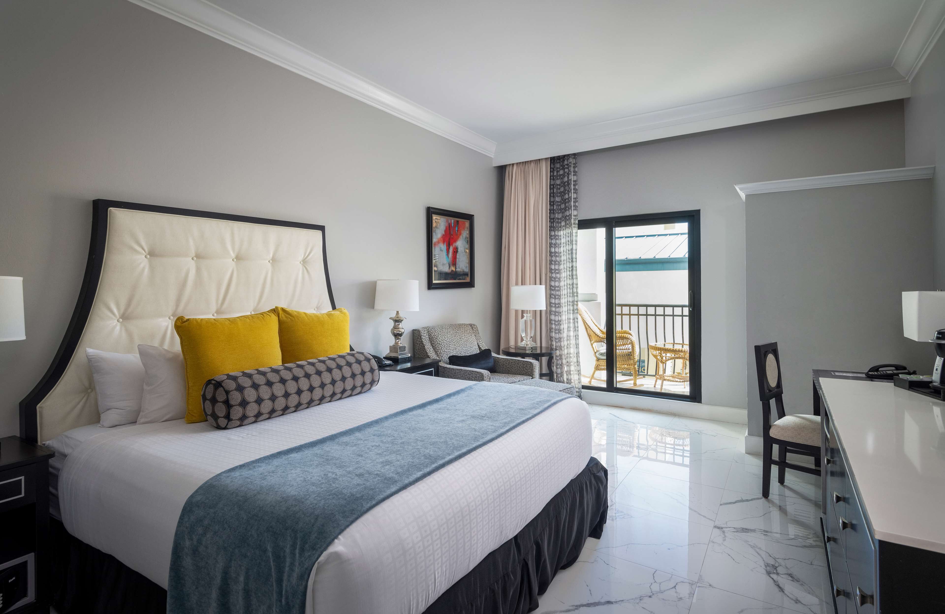 Quarto de hotel com uma cama grande, uma mesa, uma cadeira, varanda com vista e um quadro na parede.