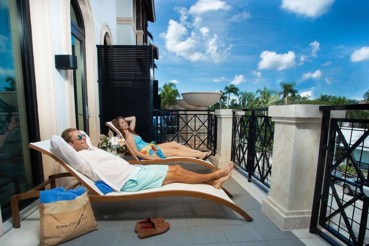 A man and a woman relaxing on lounge chairs on a balcony with a view of palm trees and a clear sky.