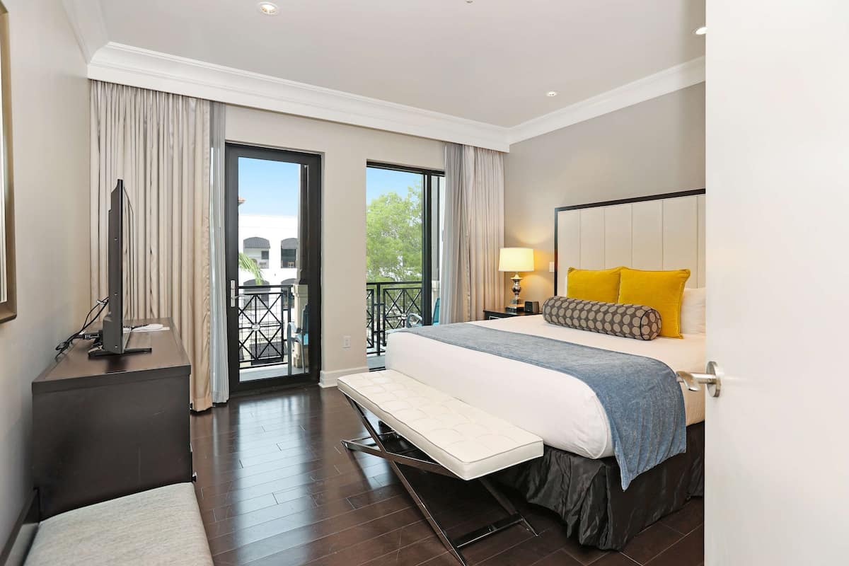 A modern hotel room with a large bed, a desk with a flat-screen TV, and a balcony with a view of greenery.
