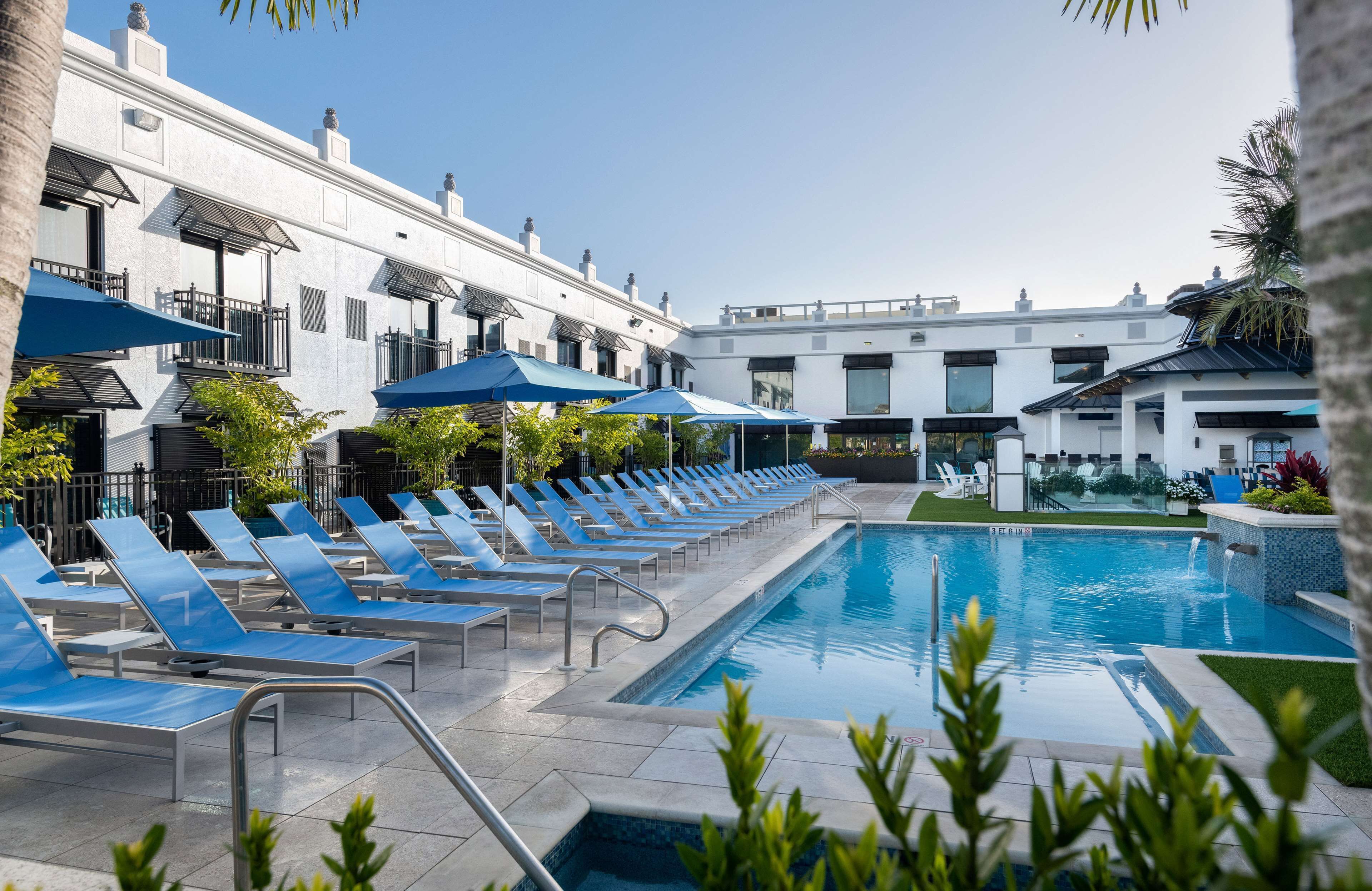 A pool area with blue lounge chairs and umbrellas, a clear sky, and a building with balconies.
