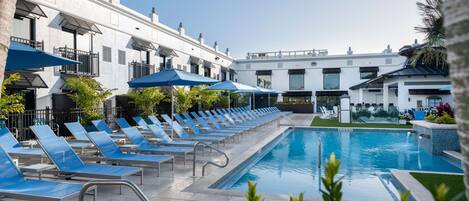 A pool area with blue lounge chairs and umbrellas, a clear sky, and a building with balconies.