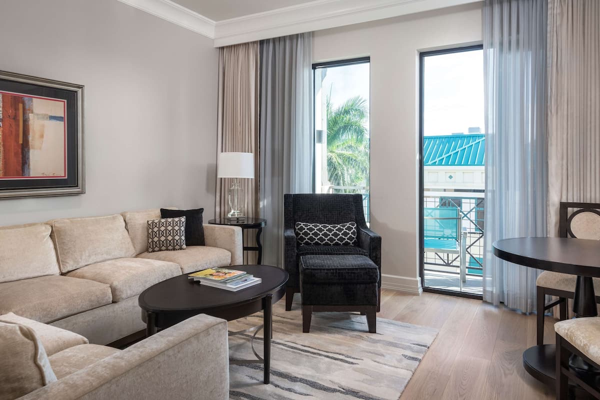 A modern hotel room with a beige sofa, a black armchair, a small round table, and a large window with a view of a blue building and palm trees.