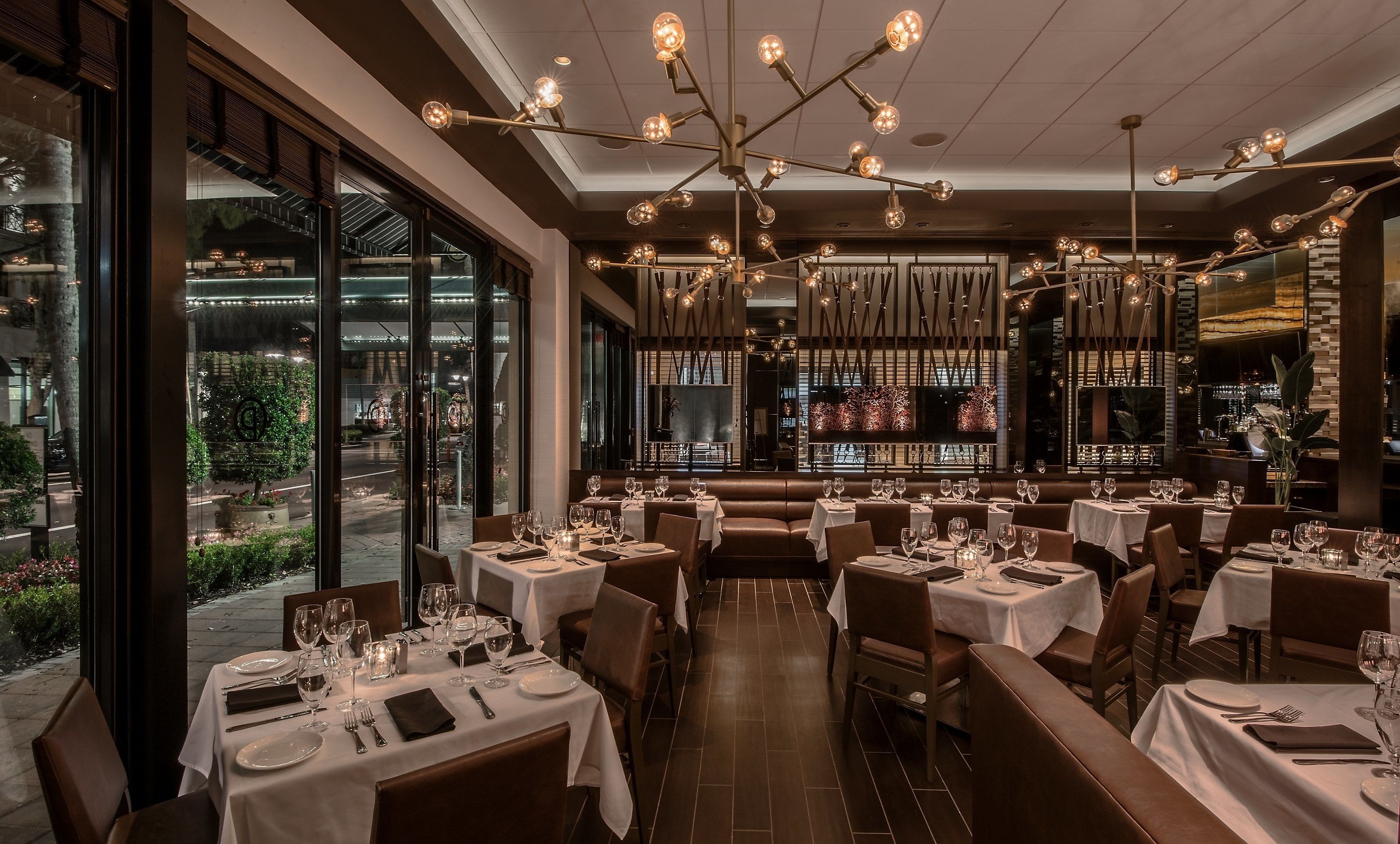 A restaurant with multiple dining tables, each set with white tablecloths, wine glasses, and cutlery. The interior features a modern chandelier and large windows offering a view of the outdoors.