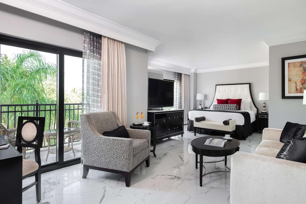 A hotel room with a bed, a sofa, a chair, a TV, and a balcony with a view of greenery.