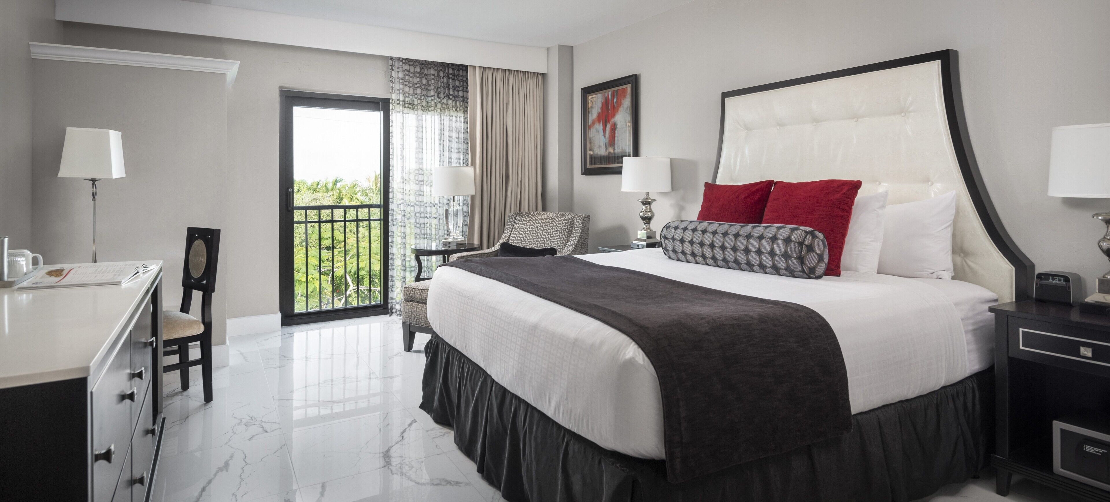 A hotel room with a large bed, a desk, a chair, a nightstand, and a view of greenery through the window.