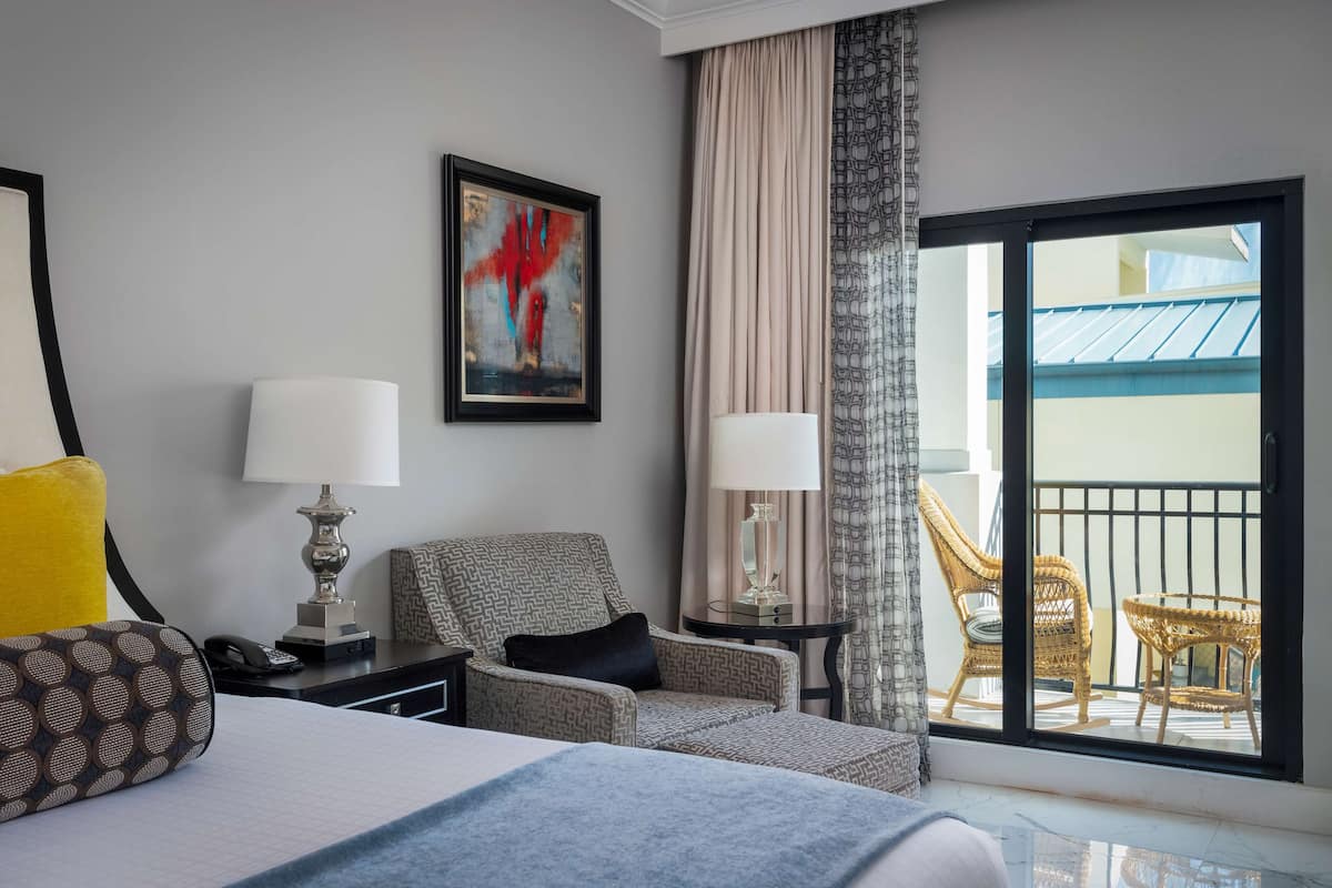A hotel room with a bed, a chair, a table with a lamp, a phone, and a balcony with wicker furniture.