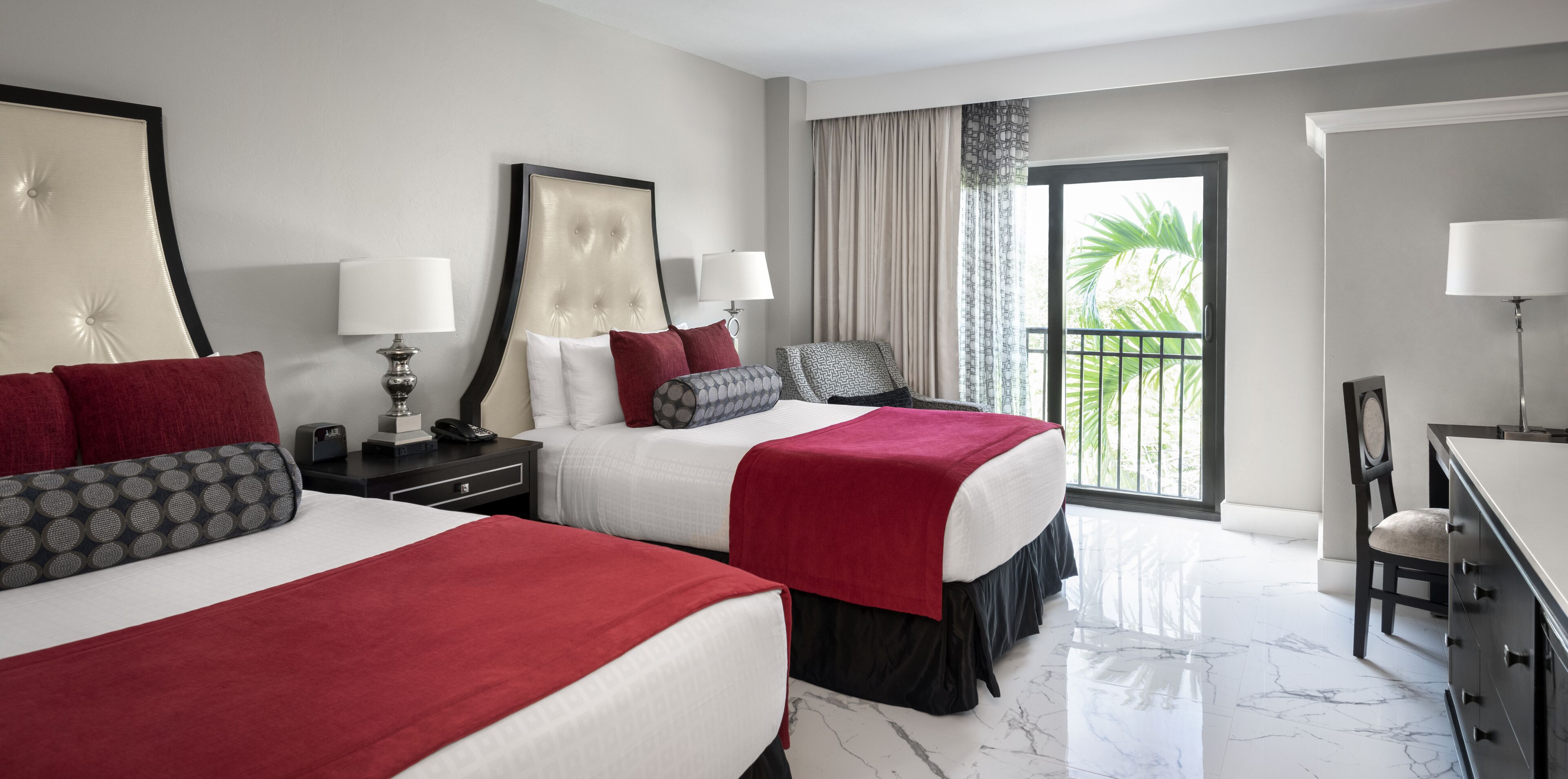 A hotel room with two beds, a desk, a chair, and a balcony with a view of greenery.