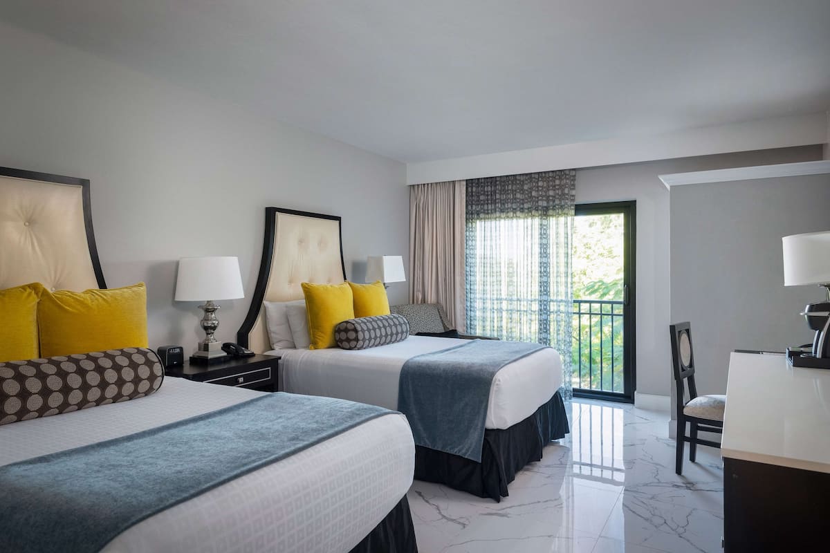 A hotel room with two beds, a desk, and a balcony with curtains.