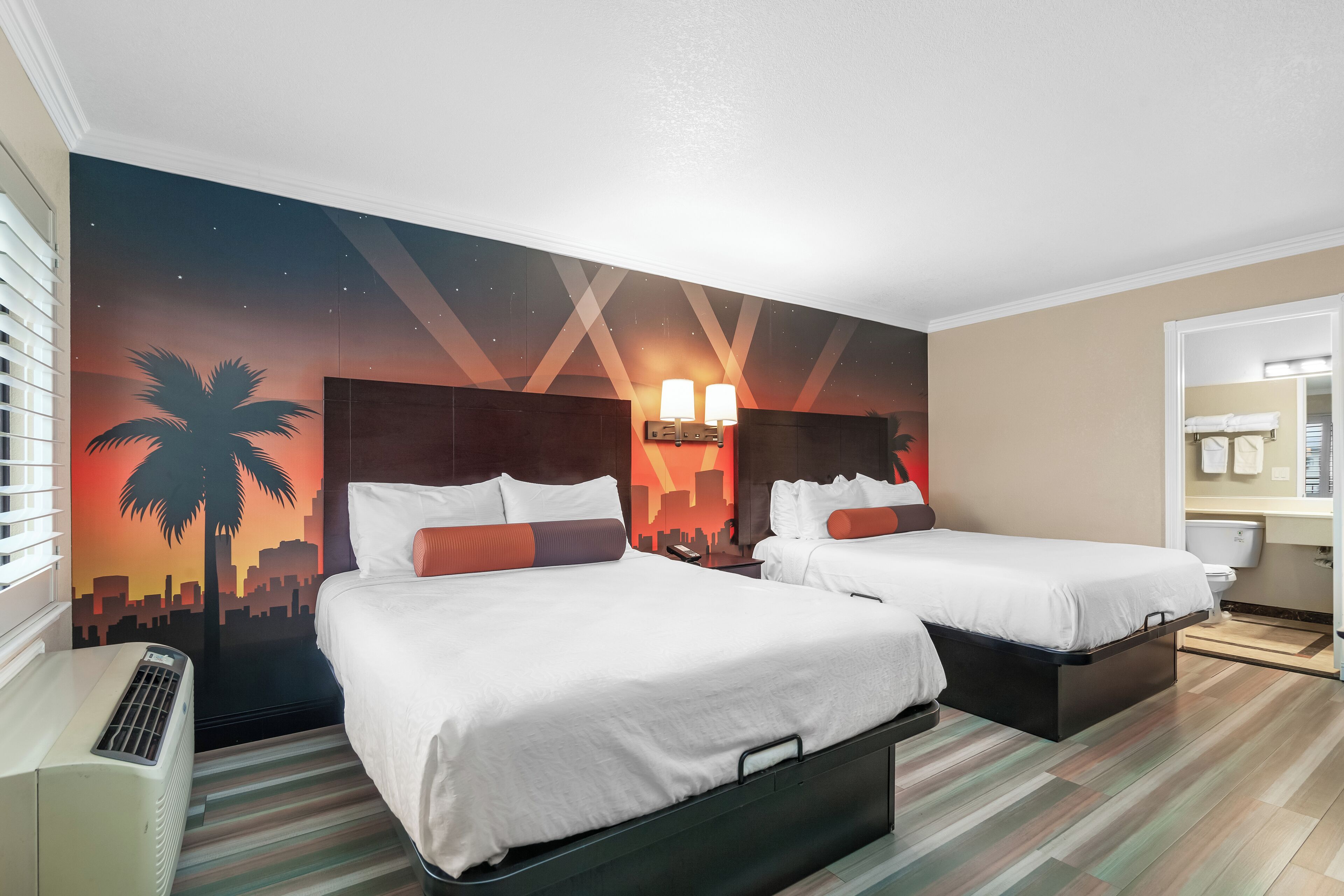 Habitación de hotel con dos camas, un mural con vista a la ciudad, un calentador y un baño con espejo y toallero.