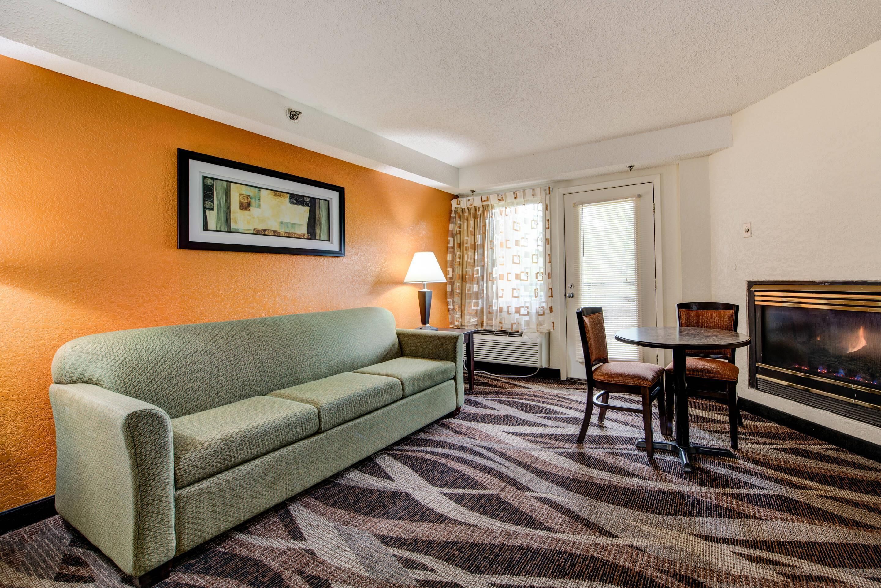 A hotel room with a green sofa, a small table with chairs, a lamp, a fireplace, and a framed picture on the wall.