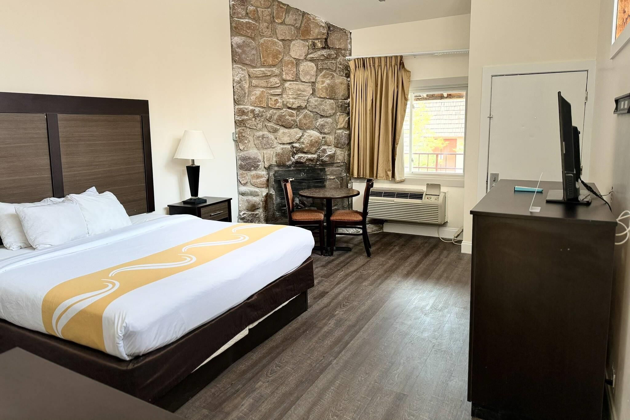 A hotel room with a bed, a stone wall, a desk with a computer, a chair, and a window with curtains.