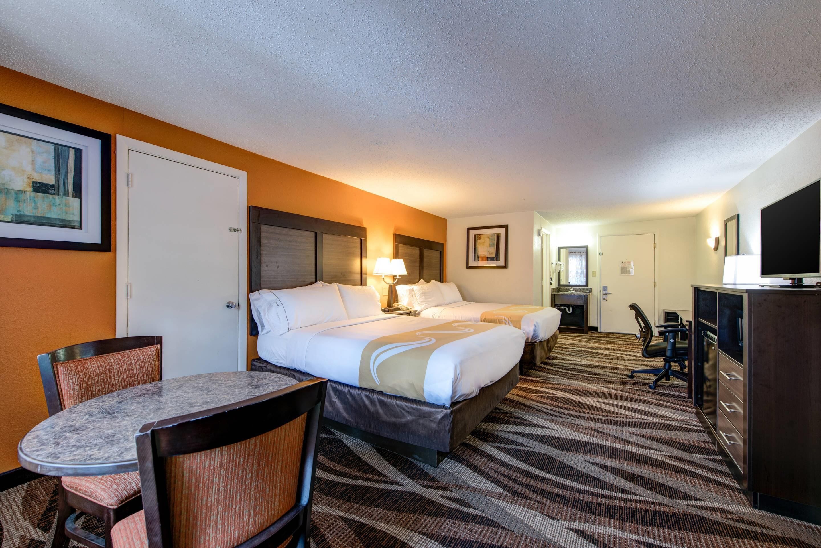 A hotel room with two beds, a dining table, a desk, and a TV.