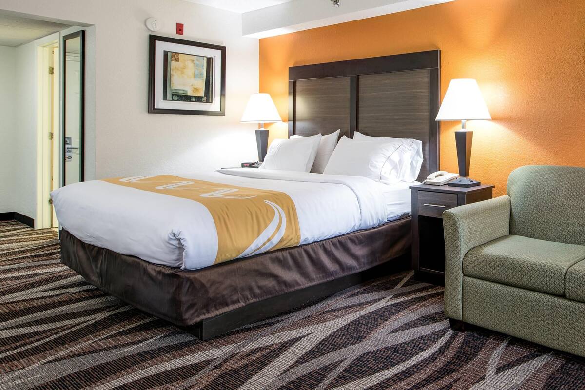 Una habitación de hotel con una cama, dos lámparas de noche, un sillón verde, un cuadro en la pared y una alfombra estampada.