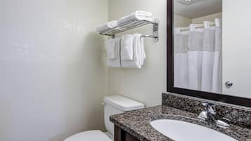 Un baño con inodoro blanco, lavamanos sobre encimera de granito, un espejo y un perchero con toallas dobladas.