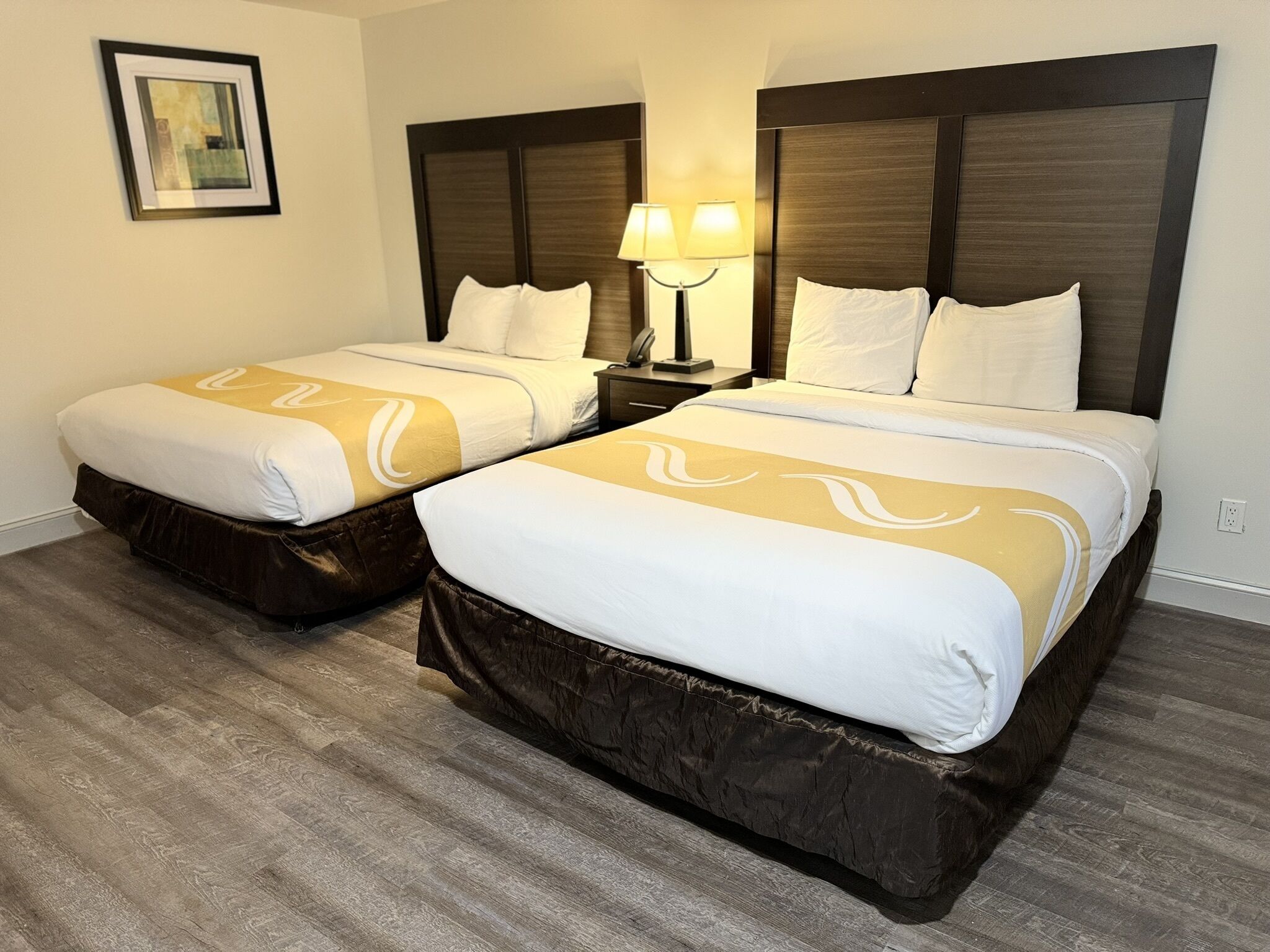 A hotel room with two beds, each with a brown headboard, a nightstand with a lamp, and a framed picture on the wall.