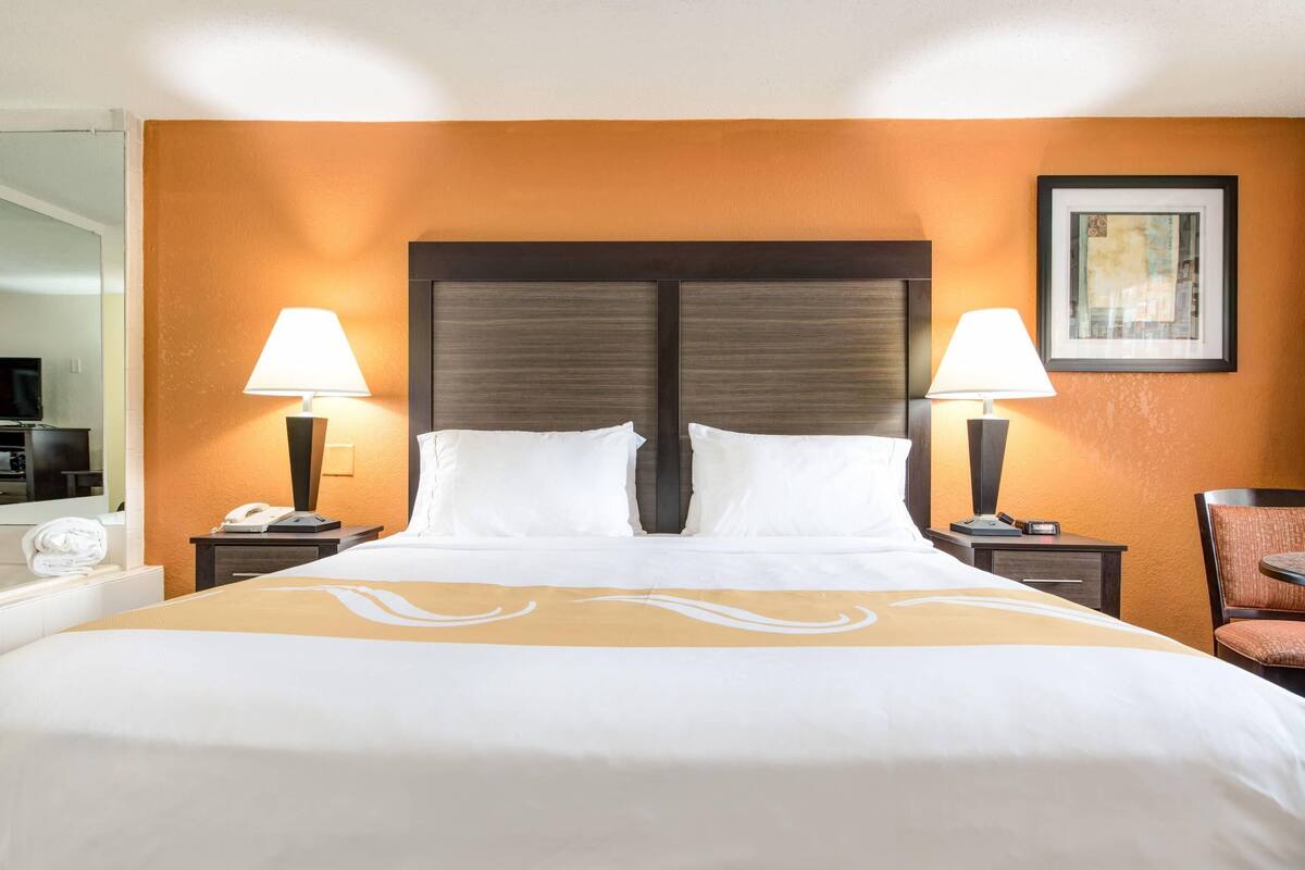 Habitación de hotel con una cama grande, dos mesitas de noche con lámparas, una silla y un cuadro en la pared.