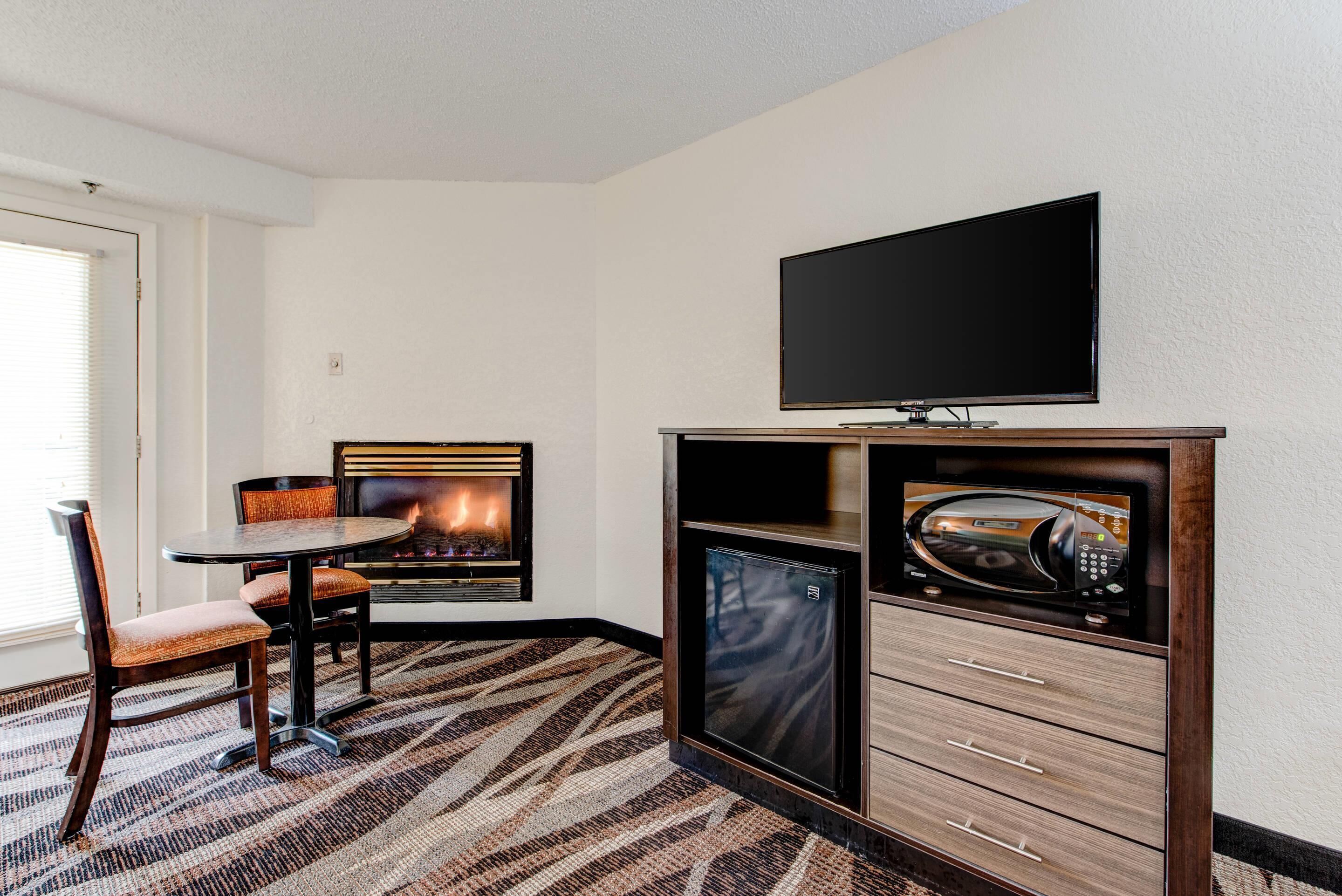 A hotel room with a fireplace, a television, a microwave, and a dining area with chairs and a table.