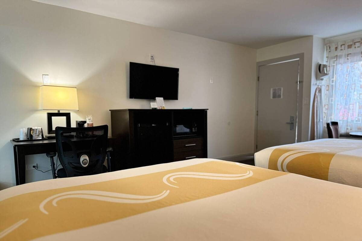 Una habitación de hotel con dos camas, un escritorio con lámpara, un televisor en la pared y una puerta.