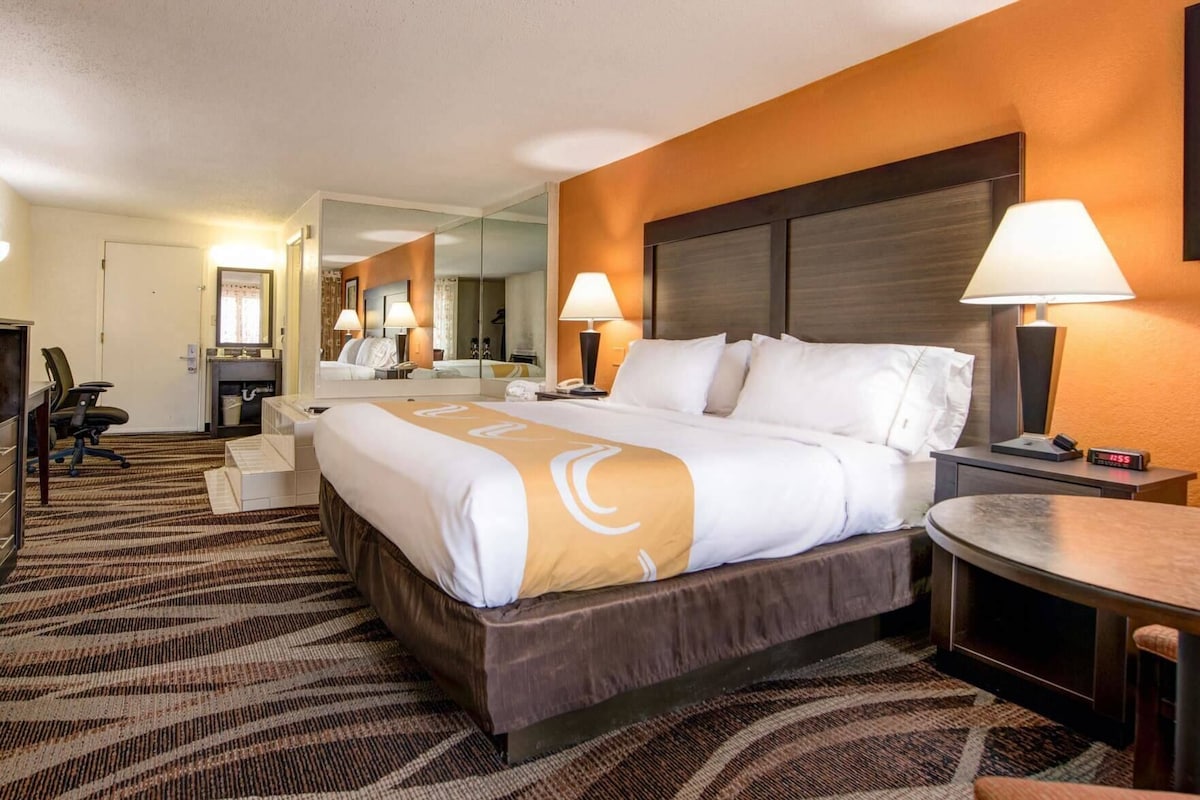 A hotel room with a large bed, bedside tables, a desk, a chair, a mirror, and a carpeted floor.