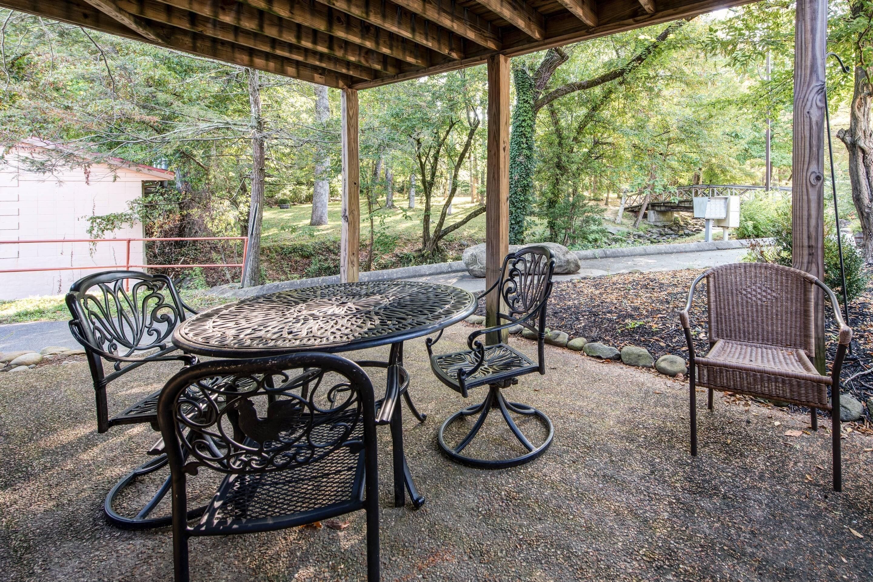 Un espace extérieur abrité, aménagé avec une table ronde en métal et quatre chaises, entouré d’arbres et bordé d’un chemin de gravier.