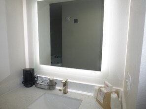 A bathroom with a large mirror, a sink, a coffee maker, and a basket containing toiletries.