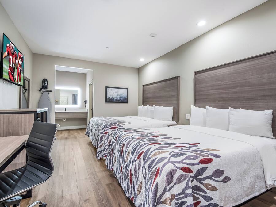 Quarto de hotel com cama, escrivaninha, cadeira, televisão e um quadro na parede.
