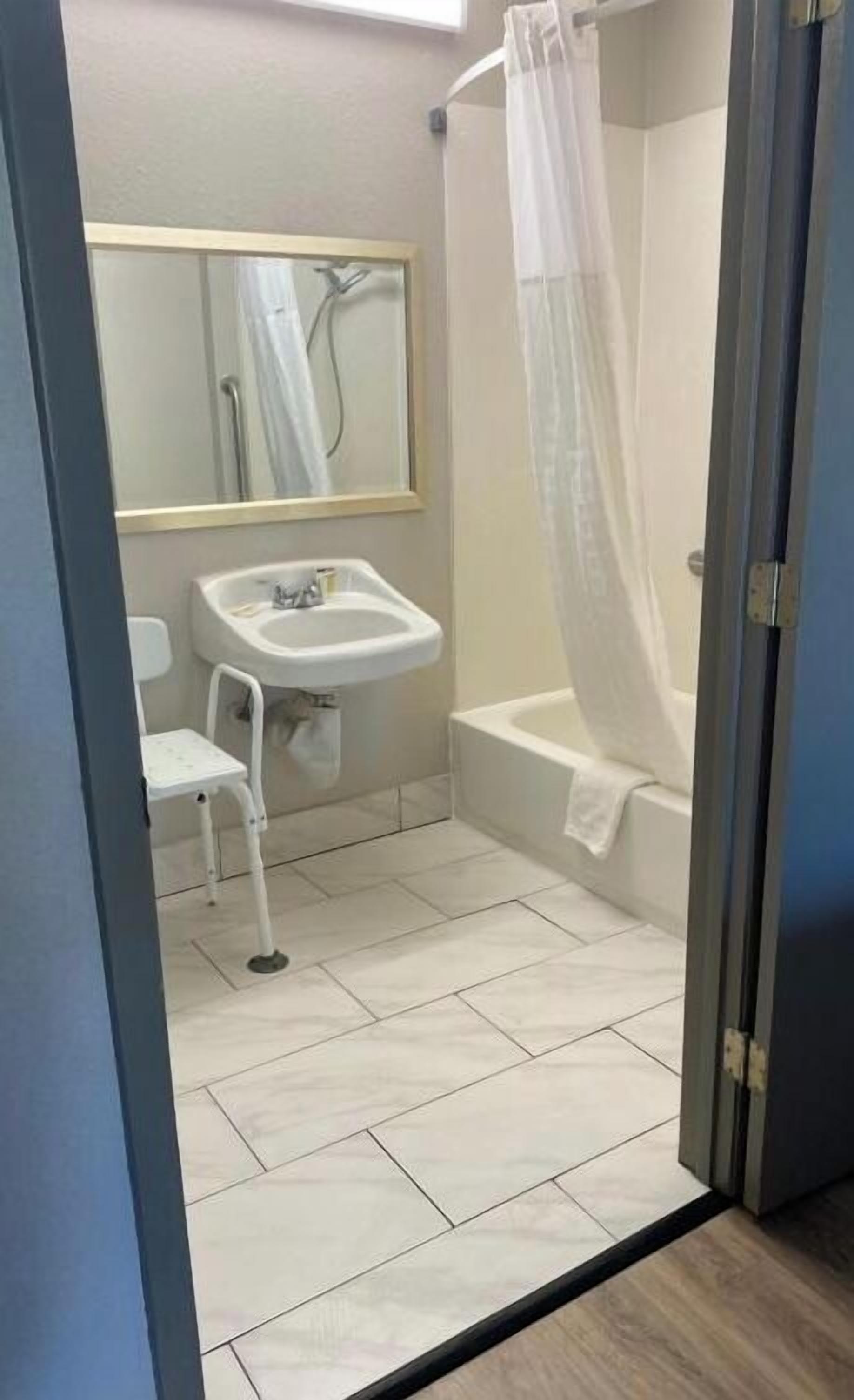 Un baño pequeño con lavamanos, espejo, ducha con cortina y una silla blanca.