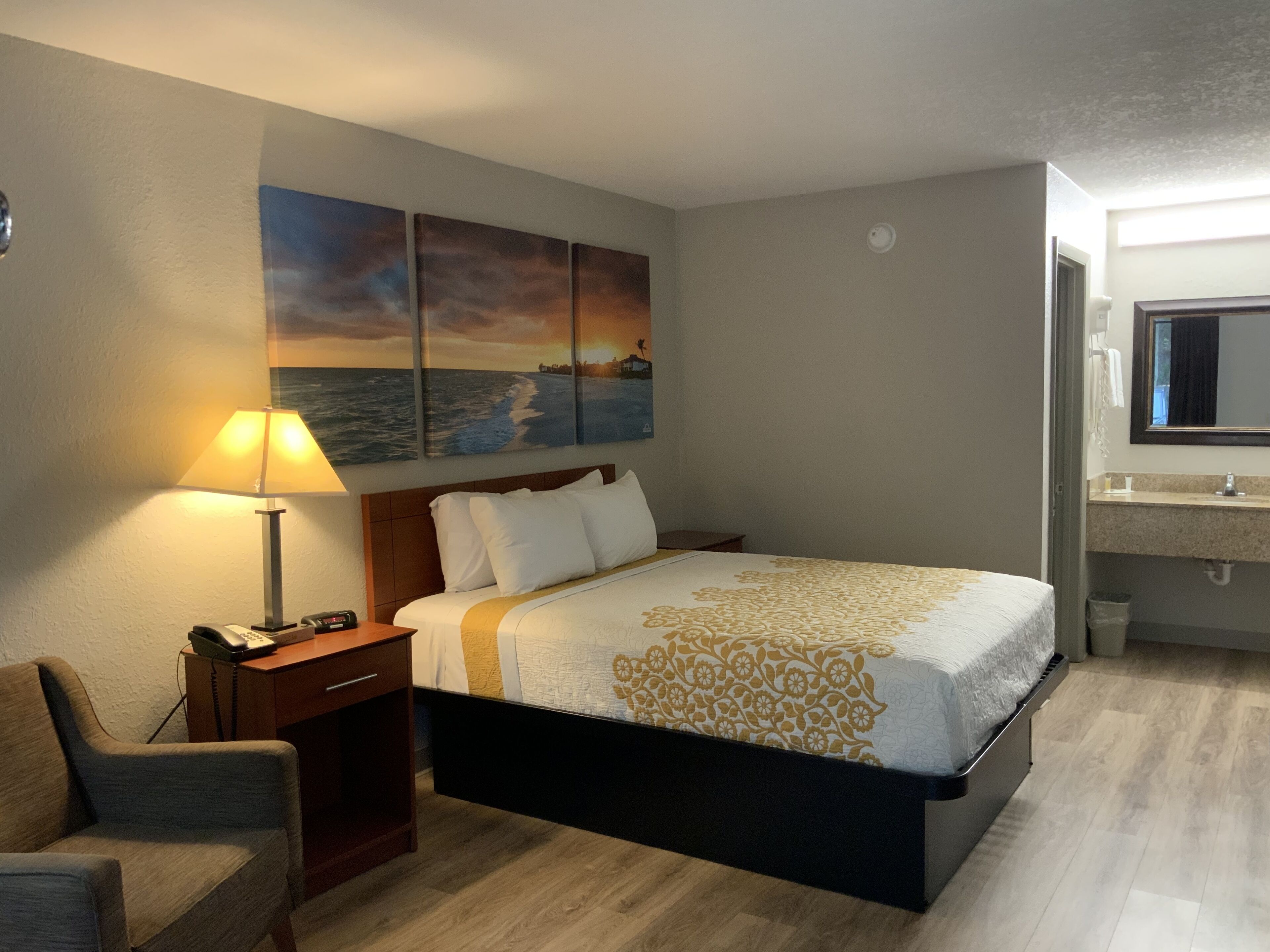 A hotel room with a bed, a desk with a lamp, a chair, a painting of a sunset over the ocean, and a bathroom with a mirror and sink.