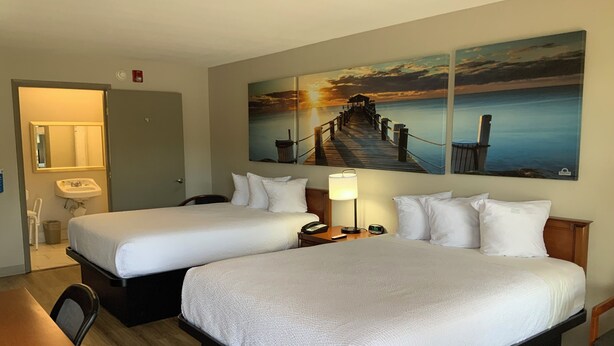 Habitación de hotel con dos camas, un escritorio, una silla, un lavamanos y un cuadro de un muelle y una puesta de sol.