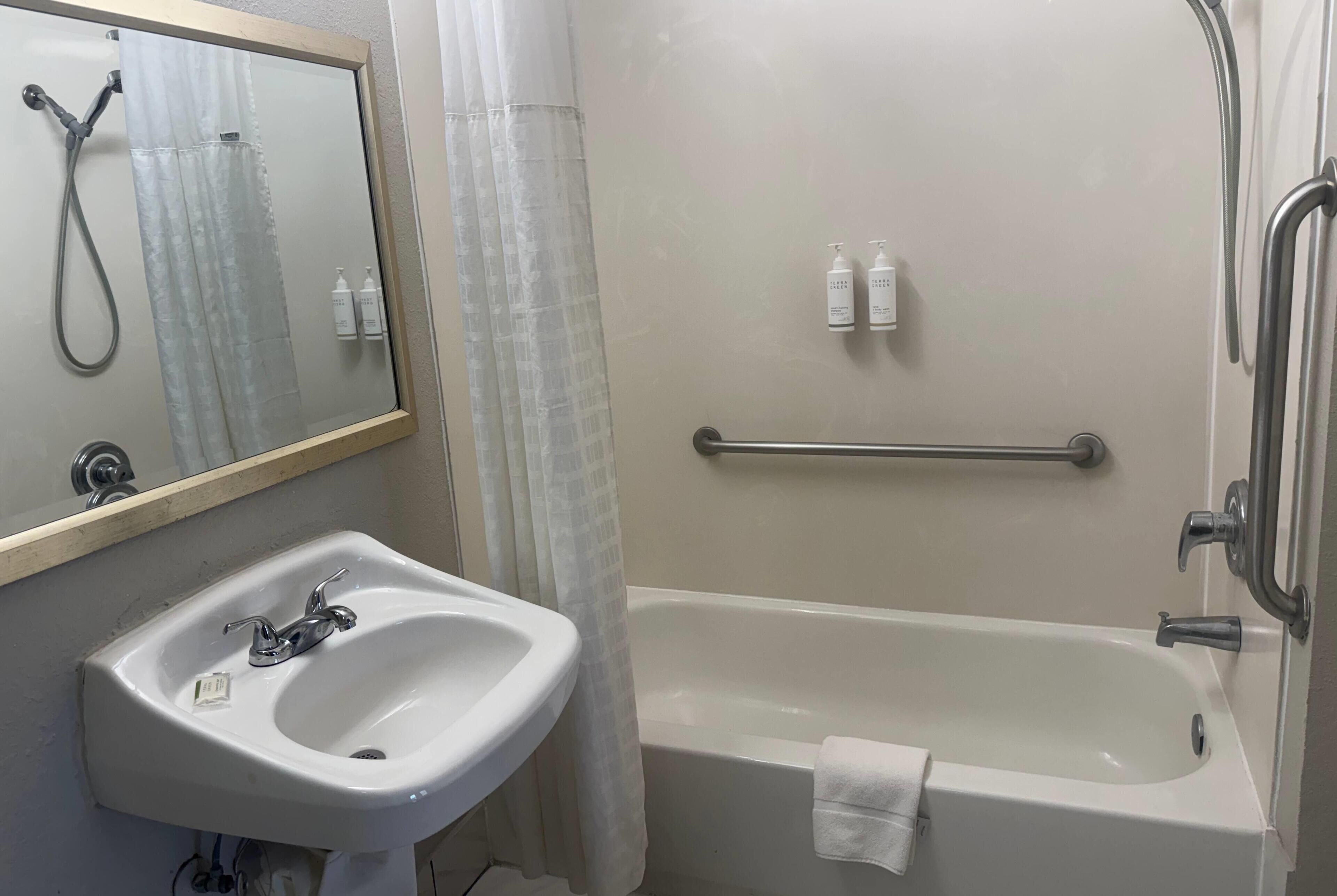A bathroom with a sink, mirror, shower, and handrails.