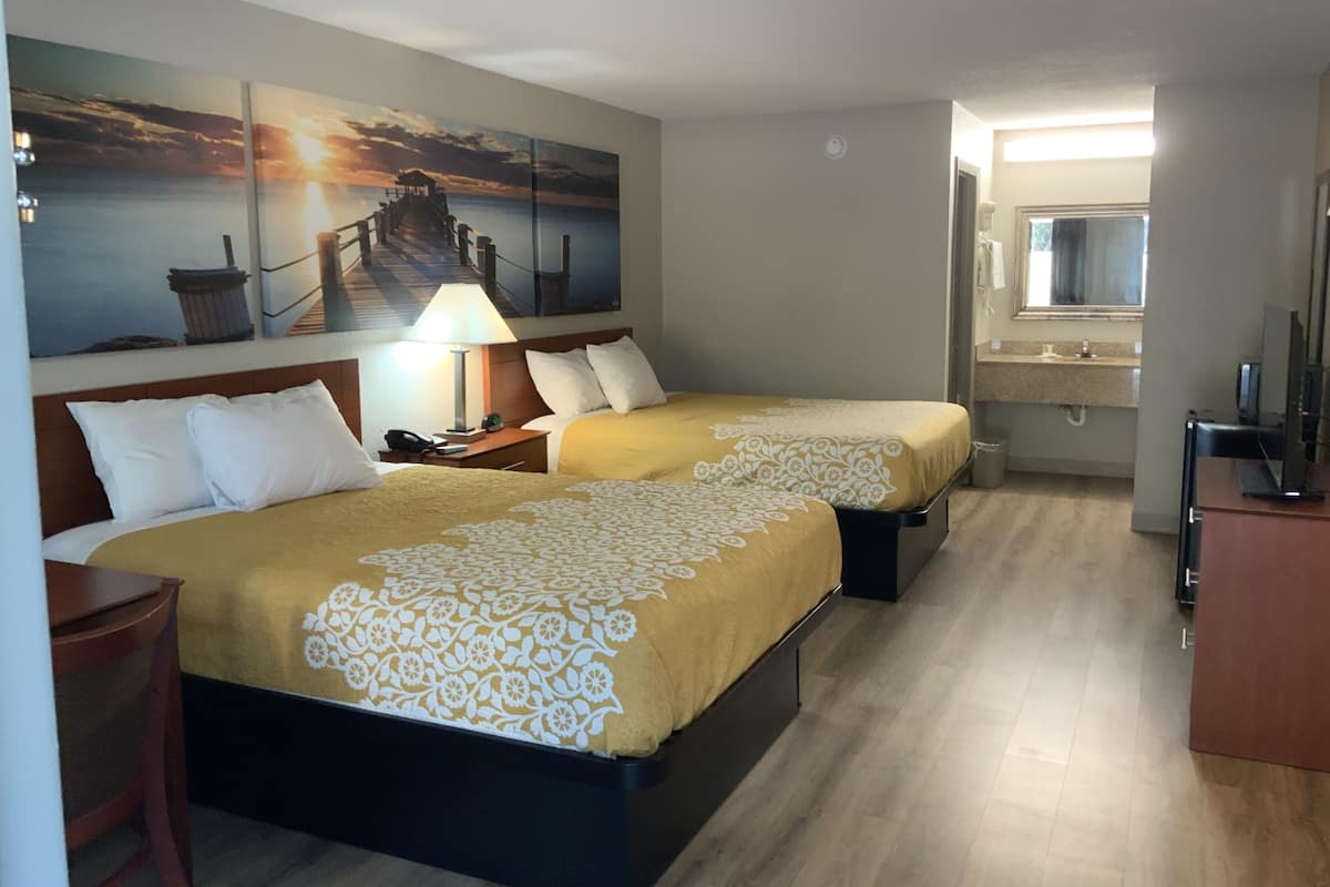 Habitación de hotel con dos camas, una mesita de noche con lámpara, un escritorio con computadora, un baño con espejo y un cuadro de un atardecer sobre un muelle.