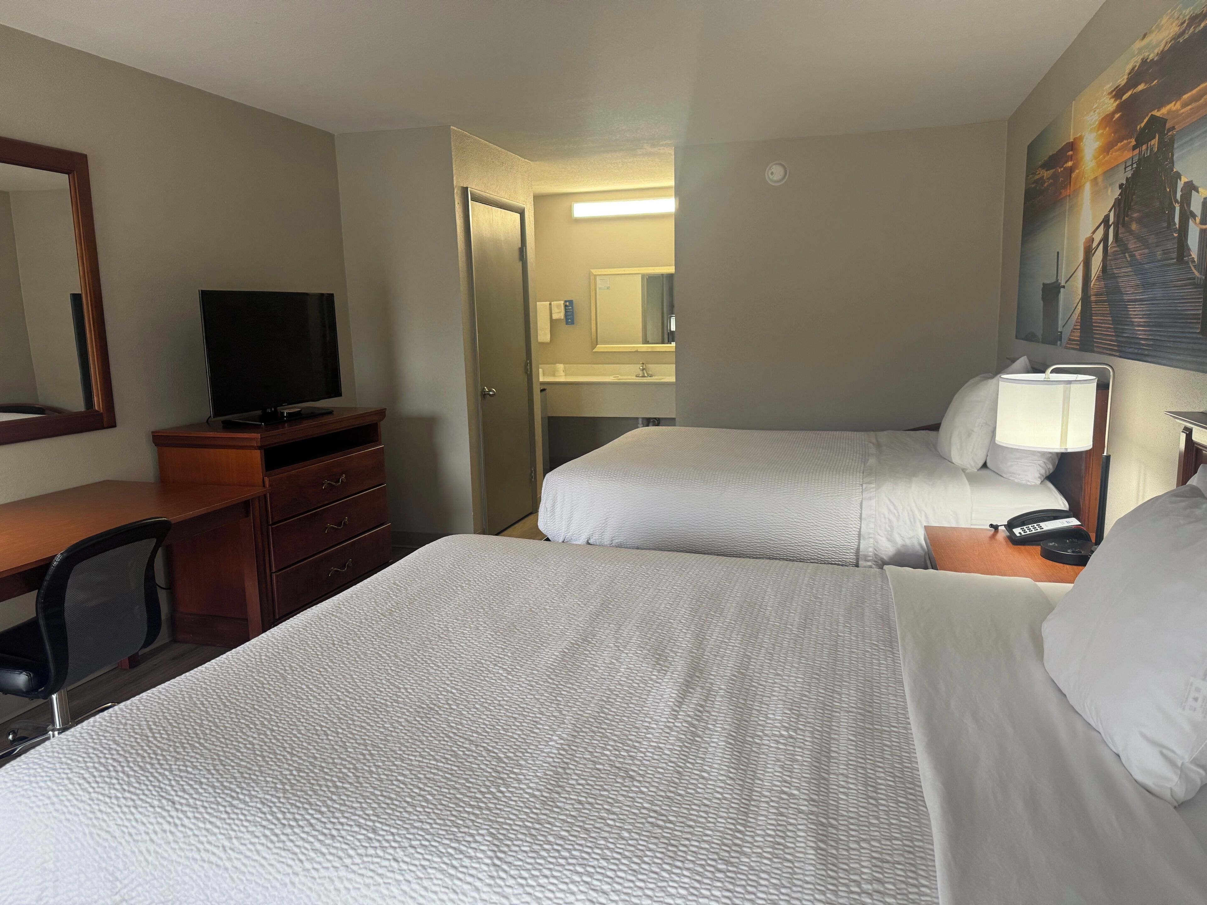 Habitación de hotel con dos camas, un escritorio con silla, un televisor, una lámpara de escritorio y un cuadro en la pared.