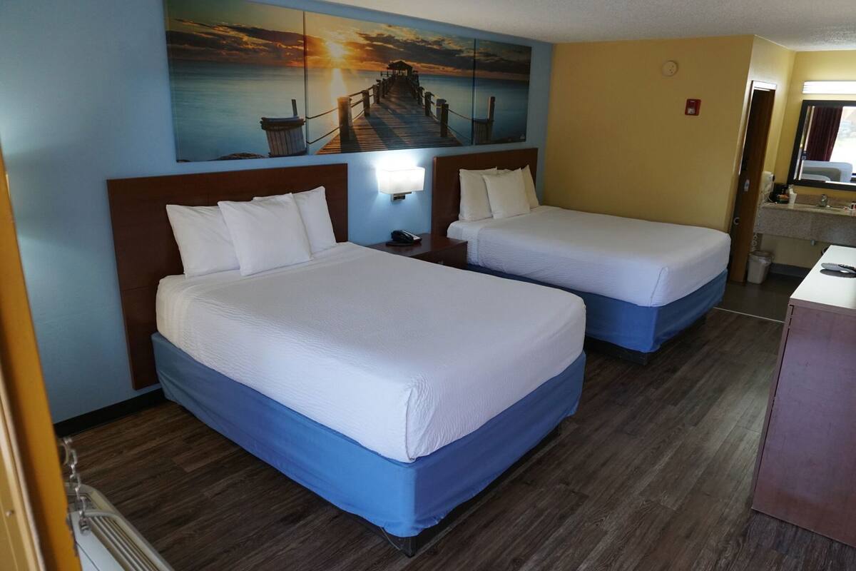 Une chambre d’hôtel avec deux lits, un tableau représentant un coucher de soleil sur l’eau, un plancher en bois et un bureau avec un miroir.