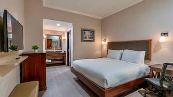 A hotel room with a large bed, a desk, a chair, a mirror, and a small plant.