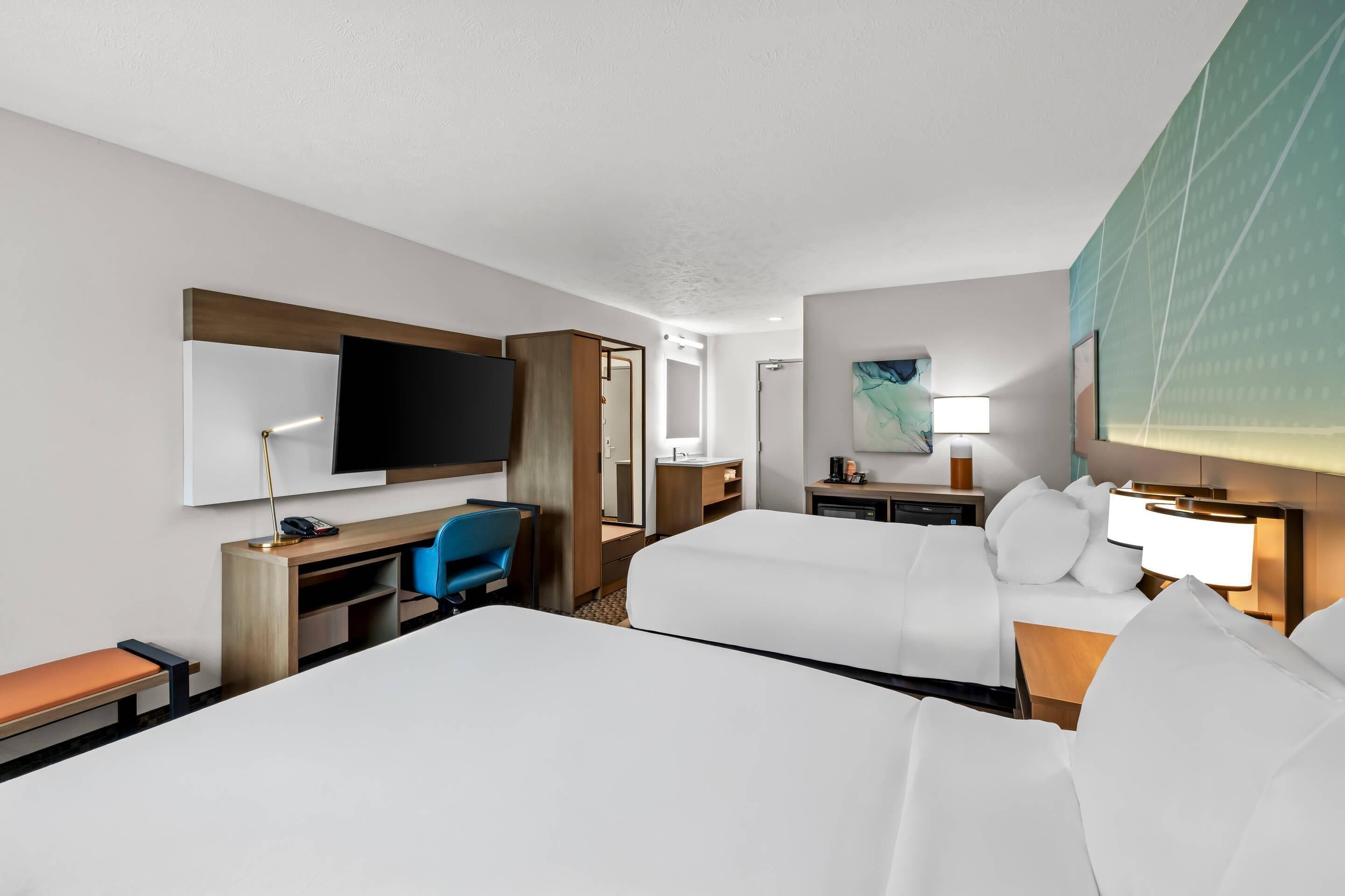 A hotel room with two beds, a desk, a chair, a TV, a desk lamp, a bedside table with a lamp, and a painting on the wall.