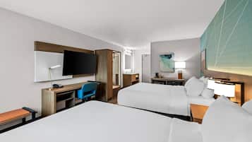 Habitación de hotel con dos camas, un escritorio, una silla, televisión, lámpara de escritorio y una mesita de noche con lámpara. Se aprecia un cuadro en la pared.