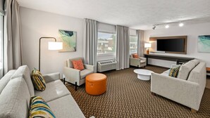 Ett modernt hotellrum med en soffa, fåtölj, pall, platt-TV och ett stort fönster med gardiner.